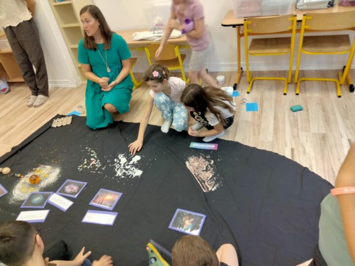 Sprievodkyňa a deti kľačia okolo čiernej plachty, na ktorej sú trblietkami, modelmi planét a kartičkami znázornené vesmír a Slnečná sústava